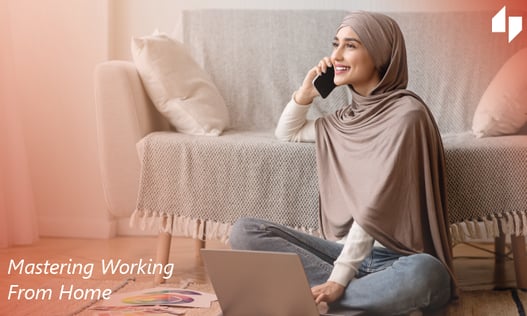 Woman sitting on the floor using a laptop and talking on the phone wearing a hijab and jeans sitting in front of a couch.